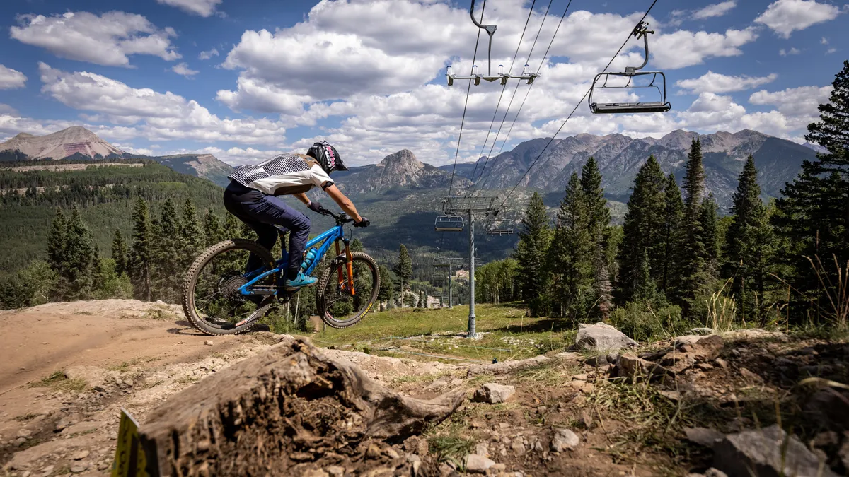 An outdoor scene featuring a mountain biker riding along a trail, with a scenic backdrop of a chairlift, expansive mountain landscape, and pine trees. The image conveys a sense of adventure and natural beauty, highlighting outdoor recreation opportunities.