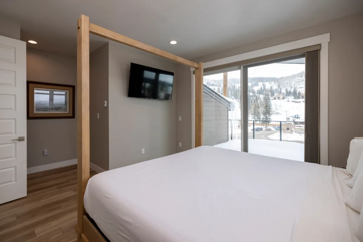 A tranquil bedroom scene with a four-poster bed, soft white linens, a mounted television, and a large window offering a breathtaking mountain view. The space feels serene and restful, ideal for both winter hibernation and cool summer nights.