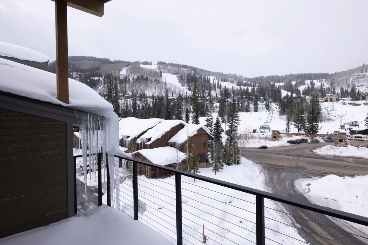 A scenic view from a balcony overlooking snow-capped mountains, icicles hanging from the roof, and rustic wooden architecture. The focus on the outdoor environment evokes the mountain serenity and the refreshing breezes enjoyed in summer.