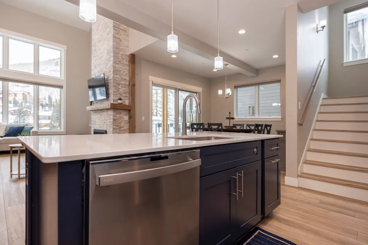A spacious, inviting interior centered on a kitchen island, with large windows letting in natural light, an adjacent dining area, and a staircase. The space suggests communal activities, family meals, and gathering spots for groups.