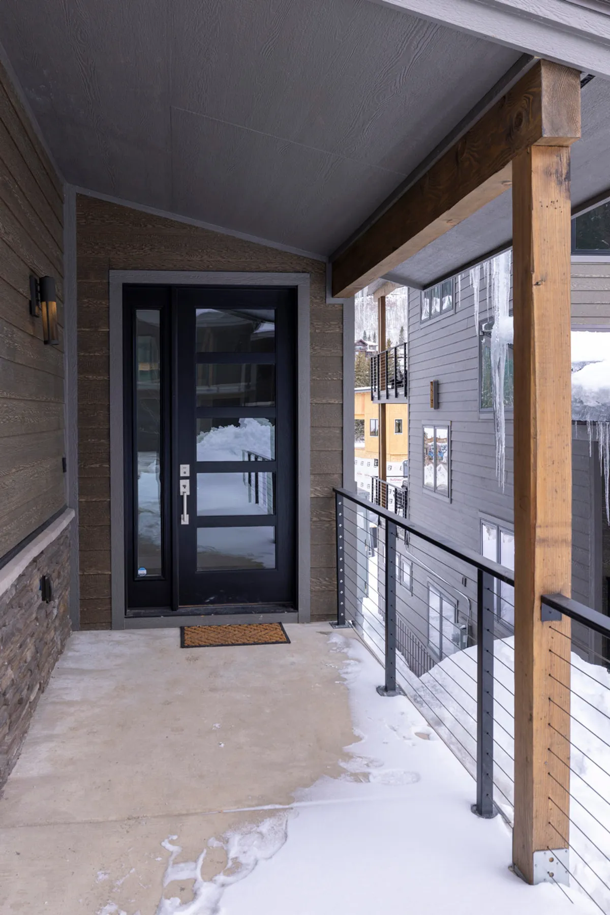 The cozy, snow-dusted front entrance of a mountain cabin, framed by a sturdy railing and surrounded by soft winter light. The scene evokes feelings of arrival and anticipation, with a welcoming atmosphere ideal for greeting guests or returning from a day on the slopes.