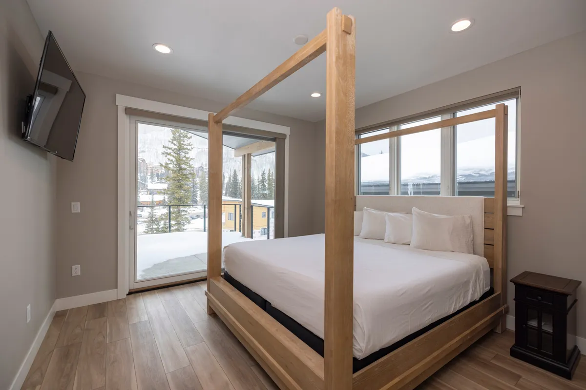 A serene interior bedroom featuring a four-poster bed dressed in luxurious white linens, complemented by large windows that bring in ample natural light. A mounted television and wooden flooring add to the comfort and elegance, creating an inviting atmosphere ideal for relaxation and restful sleep.