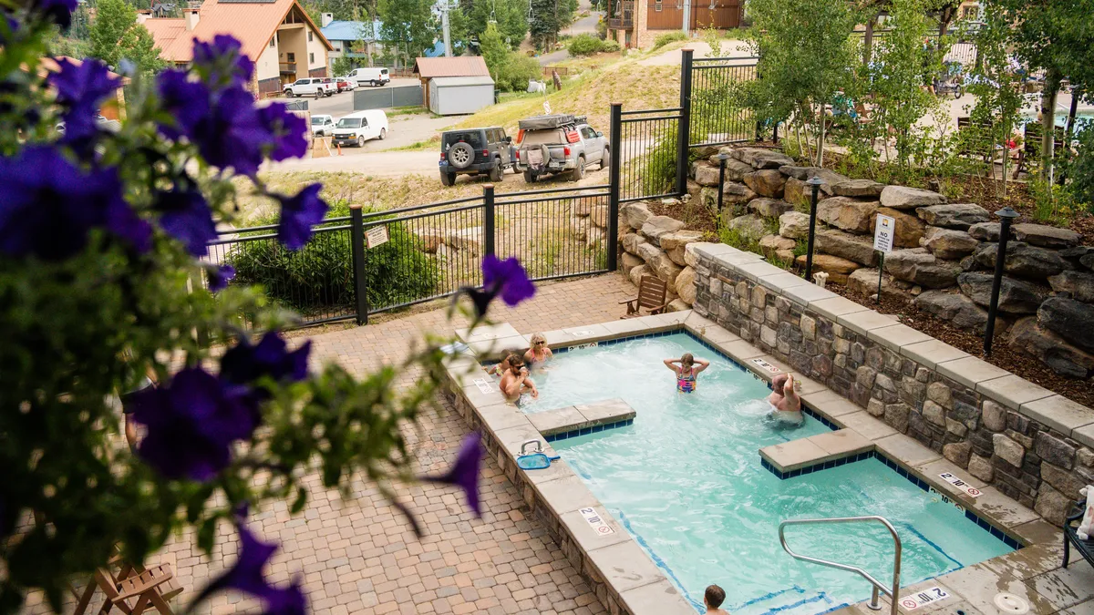 A group enjoys relaxing in an outdoor hot tub surrounded by flowers and natural landscape, with a sense of leisure and outdoor socializing. The setting feels inviting and serene, blending comfort with nature.